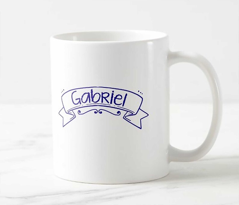 Puede incluir: Taza de cer&aacute;mica blanca con el nombre "Gabriel" en azul dentro de un dise&ntilde;o de banner. La taza tiene un asa curva y un dise&ntilde;o simple y limpio. El texto es de estilo manuscrito.