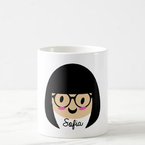 May include: White ceramic mug featuring a cartoon illustration of a person with black hair, glasses, and pink cheeks. The name "Sofia" is written in cursive below the face. The mug is cylindrical and sits on a white surface.