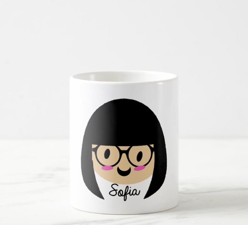 May include: White ceramic mug featuring a cartoon illustration of a person with black hair, glasses, and pink cheeks. The name "Sofia" is written in cursive below the face. The mug is cylindrical and sits on a white surface.