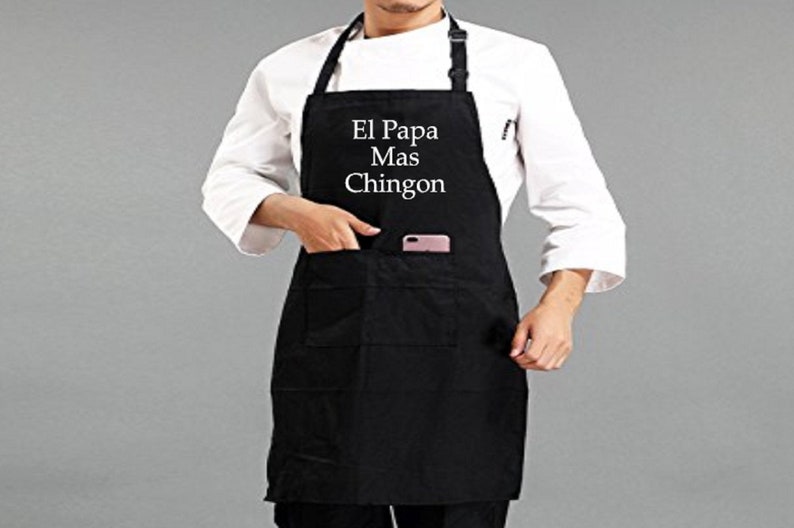Pu&ograve; includere: Grembiule nero con la scritta bianca "El Papa Mas Chingon". Il grembiule ha una tasca e un cinturino regolabile per il collo. Un telefono &egrave; visibile nella tasca. La persona indossa una giacca da chef bianca.