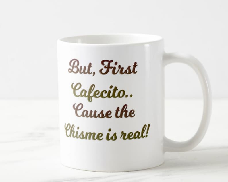 Może przedstawiać: Biały ceramiczny kubek z napisem "But, First Cafecito.. Cause the Chisme is real!" w brązowo-złotych literach. Kubek ma zakrzywiony uchwyt.