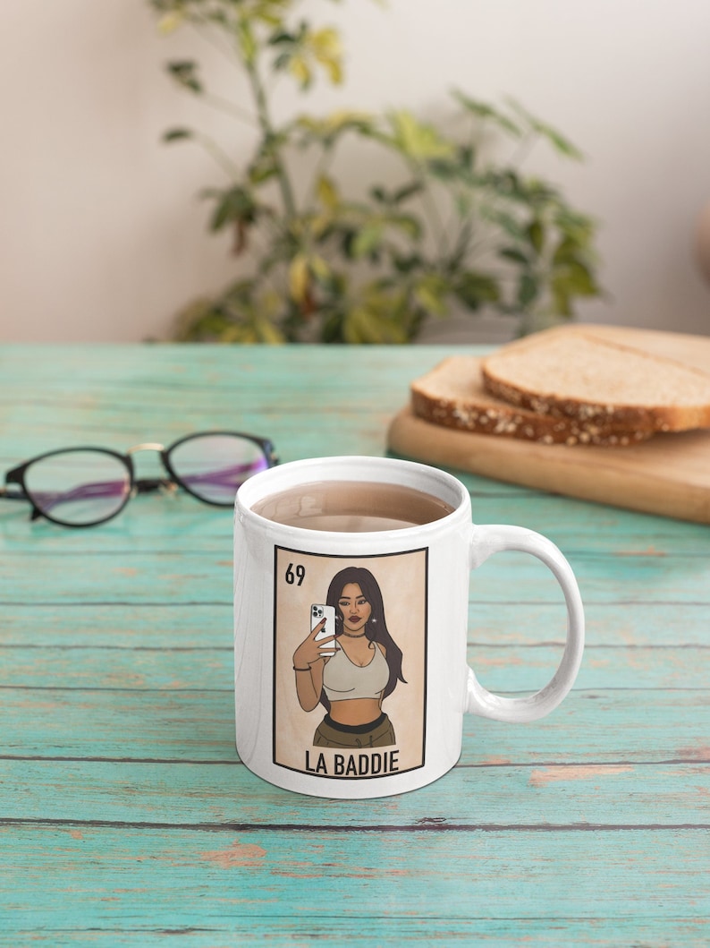 Pu&ograve; includere: Tazza in ceramica bianca con un'illustrazione colorata di una donna che si fa un selfie, con la scritta "LA BADDIE" sotto. La tazza ha un manico ed &egrave; riempita di liquido. Il numero "69" si trova nell'angolo in alto a sinistra dell'illustrazione.