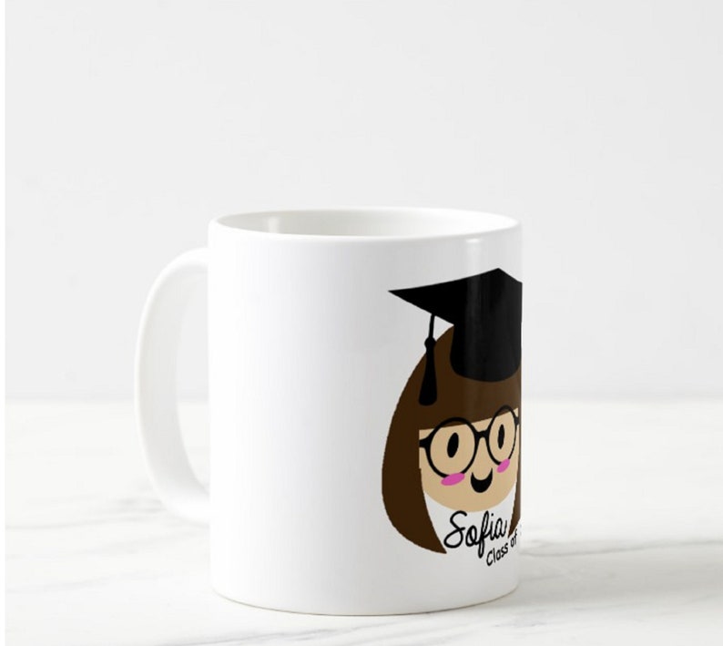 May include: White ceramic mug featuring a cartoon illustration of a graduate with brown hair, glasses, and a black graduation cap. The mug has the name "Sofia" and "Class of" printed on it.