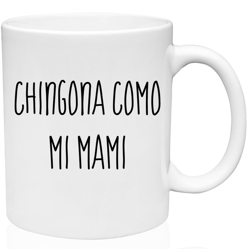 May include: White ceramic mug with the text "CHINGONA COMO MI MAMI" in black, handwritten-style lettering. The mug has a standard handle and a glossy finish. A simple, everyday coffee or tea mug.