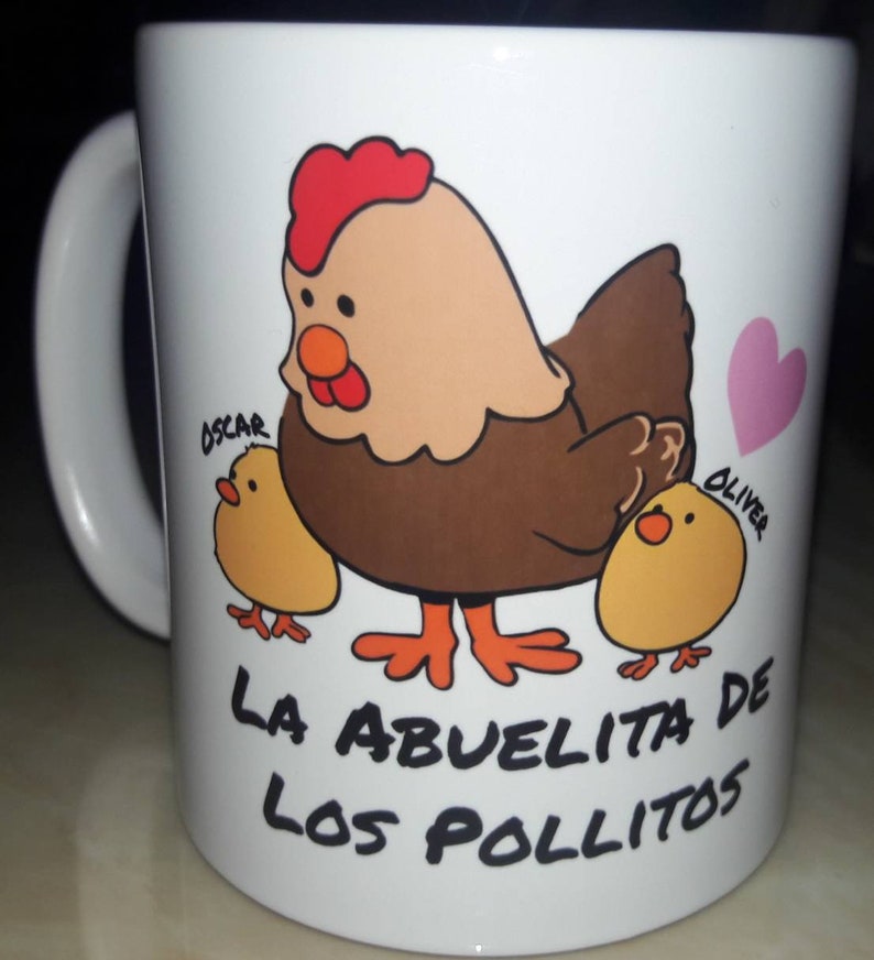 May include: White ceramic mug featuring a cartoon hen with two chicks and a pink heart. The hen is brown, tan, and red, with the text "LA ABUELITA DE LOS POLLITOS" below the image. The chicks are yellow, and the names "OSCAR" and "OLIVER" are written on the mug.