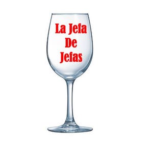 May include: A clear wine glass with a stem. The words "La Jefa De Jefas" are printed in red on the glass. The glass is empty and sits on a white background.