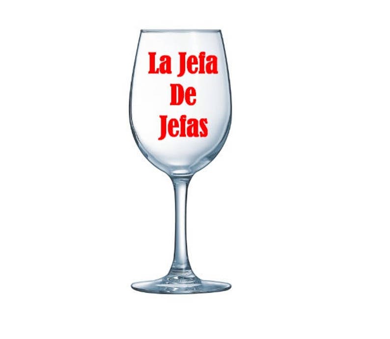 May include: A clear wine glass with a stem. The words "La Jefa De Jefas" are printed in red on the glass. The glass is empty and sits on a white background.