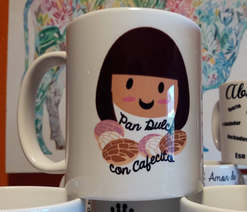 May include: White ceramic mug featuring a cartoon girl with brown hair and a smiling face. The mug has the text "Pan Dulce con Cafecito" and illustrations of sweet bread. A handle is on the left side of the mug.