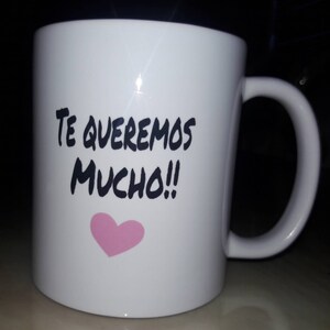 May include: White ceramic mug with a curved handle. The mug features the text "TE QUEREMOS MUCHO!!" in black, with a pink heart below the text. The mug is designed for hot beverages.