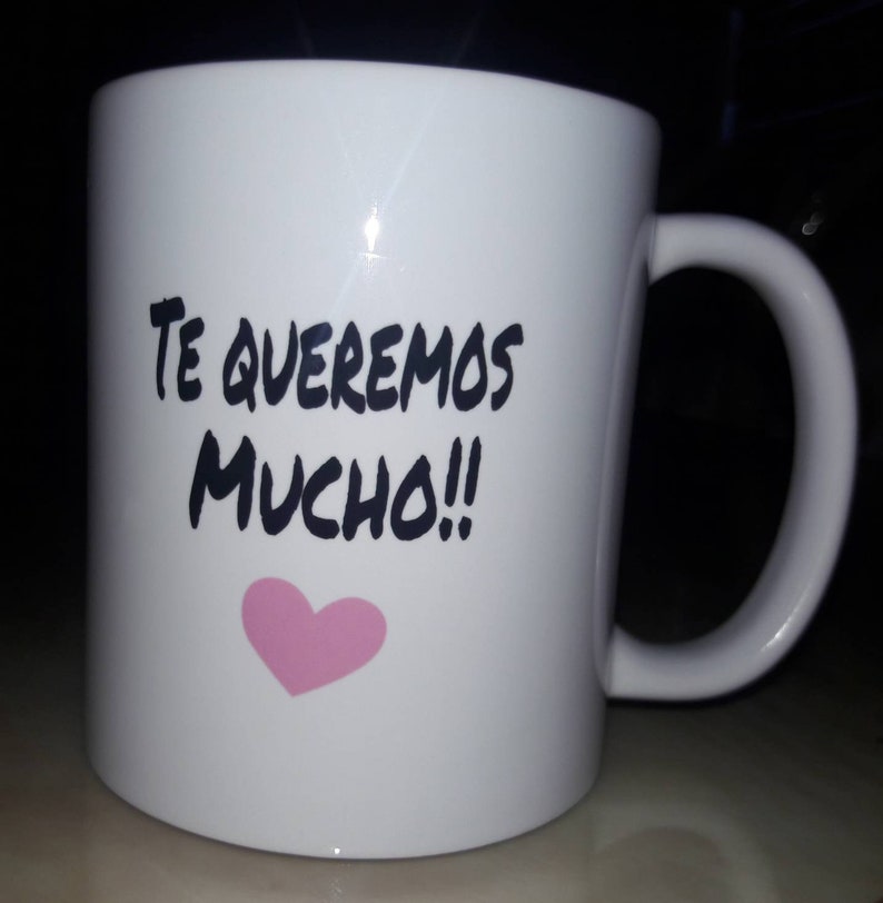 May include: White ceramic mug with a curved handle. The mug features the text "TE QUEREMOS MUCHO!!" in black, with a pink heart below the text. The mug is designed for hot beverages.
