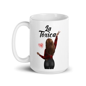 以下が含まれることがあります： 白いセラミックマグカップ。長い茶色の髪の人が赤いチェックのシャツと黒いパンツを着ているグラフィックが描かれています。マグカップには「La Toxica」の文字とピンクのハートが描かれています。