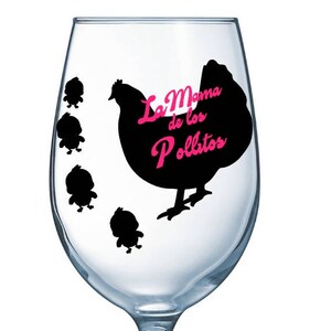 May include: A clear wine glass with a black hen and chick design. The hen has the text "La Mama de los Pollitos" in pink. Four small black chicks are to the left of the hen. The glass is empty.