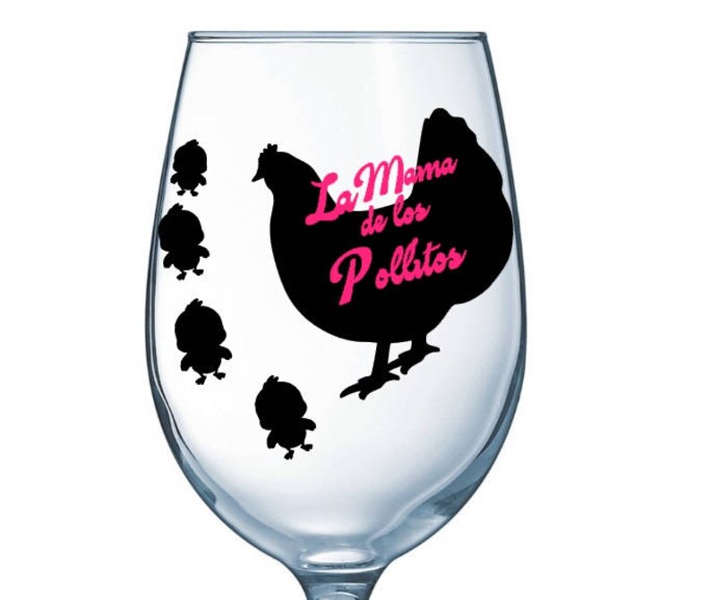 May include: A clear wine glass with a black hen and chick design. The hen has the text "La Mama de los Pollitos" in pink. Four small black chicks are to the left of the hen. The glass is empty.