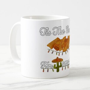 Może przedstawiać: Biały ceramiczny kubek z świątecznym wzorem. Kubek zawiera napis "Tis The Season For Tamales" z ilustracjami tamales, kaktusa w sombrero i łańcuch&oacute;w świetlnych.