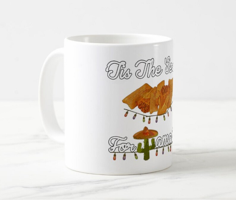 Może przedstawiać: Biały ceramiczny kubek z świątecznym wzorem. Kubek zawiera napis "Tis The Season For Tamales" z ilustracjami tamales, kaktusa w sombrero i łańcuch&oacute;w świetlnych.