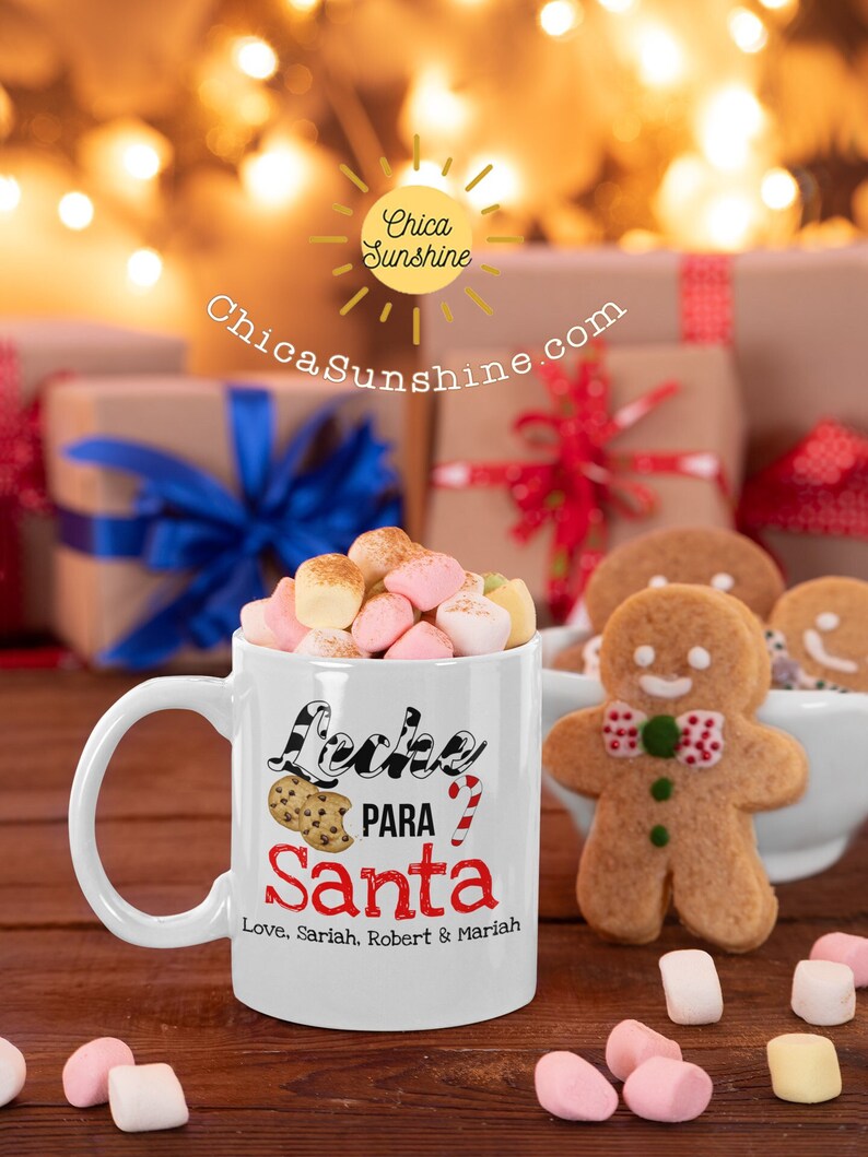 May include: A white mug filled with marshmallows, with the text "Leche Para Santa" and a cookie and candy cane graphic. A gingerbread man and gifts are in the background. The mug says "Love, Sariah, Robert & Mariah".