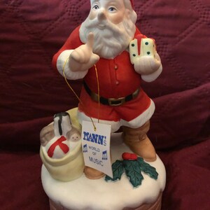 May include: A ceramic Santa Claus figurine in a red suit with a black belt, holding a gift and pointing. A tag reads "MANN'S WORLD of MUSIC". The figurine stands on a snowy chimney base with a sack of gifts.