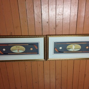 May include: Two framed golf-themed prints. Each features a dark blue background with golf clubs, a golf ball, and an oval image of a golfer. The frames are gold-coloured with a decorative edge.