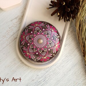 May include: A round, brown stone painted with a pink and gold mandala design. The stone is on a white tray with a brown pine branch in the background.