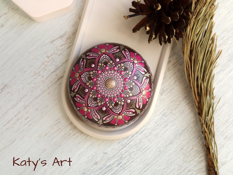 May include: A round, brown stone painted with a pink and gold mandala design. The stone is on a white tray with a brown pine branch in the background.