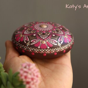 May include: A hand holds a round, brown stone painted with a pink and gold mandala design. The mandala features intricate details and a central dot.