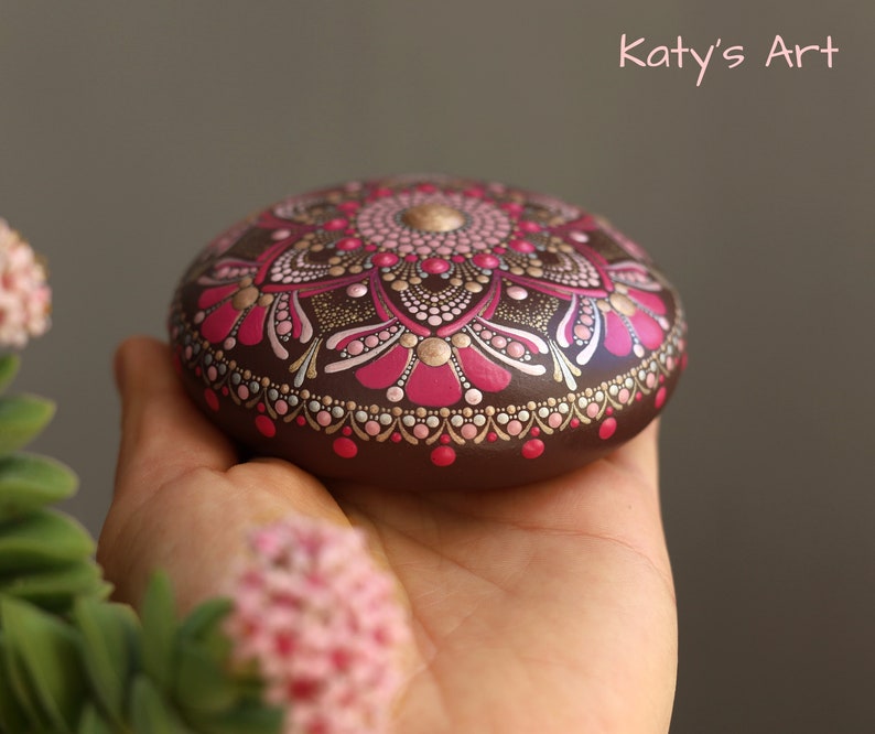 May include: A hand holds a round, brown stone painted with a pink and gold mandala design. The mandala features intricate details and a central dot.