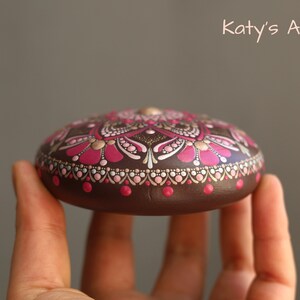 May include: A hand holds a brown stone painted with a pink and gold mandala design. The stone is round and has a smooth surface. "Katy's Art" is written in white text on a gray background.