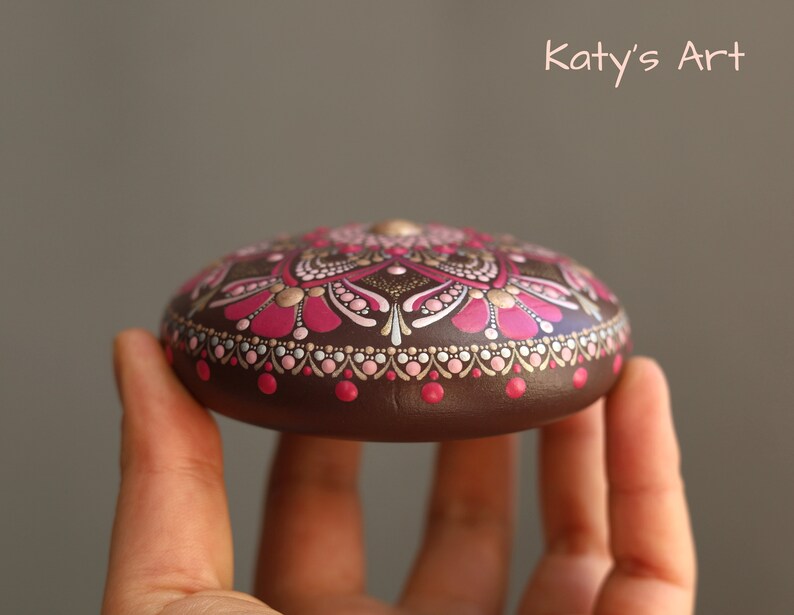 May include: A hand holds a brown stone painted with a pink and gold mandala design. The stone is round and has a smooth surface. "Katy's Art" is written in white text on a gray background.