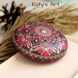 May include: A brown stone painted with a pink, gold, and white mandala design. The stone is on a light brown tray.