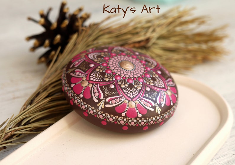 May include: A brown stone painted with a pink, gold, and white mandala design. The stone is on a light brown tray.
