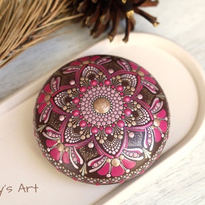 May include: A round, brown stone painted with a pink and gold mandala design. The stone is on a white plate. "Katy's Art" is written in black text at the bottom of the image.
