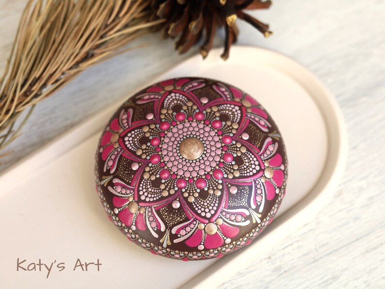 May include: A round, brown stone painted with a pink and gold mandala design. The stone is on a white plate. "Katy's Art" is written in black text at the bottom of the image.