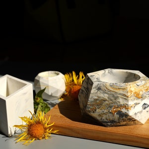 May include: Three geometric concrete planters in white, grey, and gold with a marbled effect. The planters are on a wooden surface with yellow flowers.