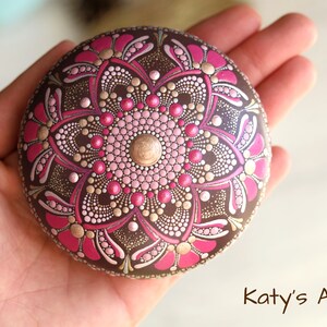 May include: A round, brown stone painted with a pink and gold mandala design. The mandala features a central flower with intricate details and a gold dot in the center.