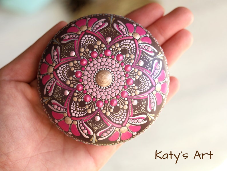 May include: A round, brown stone painted with a pink and gold mandala design. The mandala features a central flower with intricate details and a gold dot in the center.