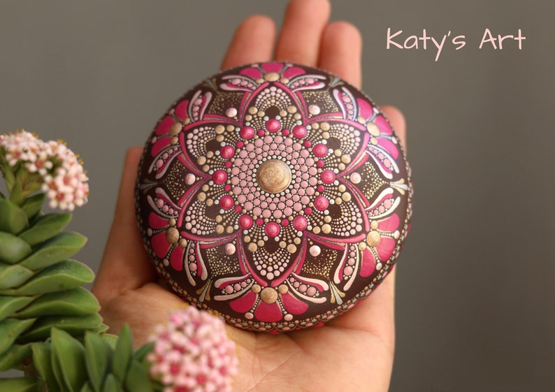 May include: A hand holds a round, brown stone painted with a pink and gold mandala design. The stone is decorated with intricate details and dots. The text "Katy's Art" is visible in the upper right corner of the image.