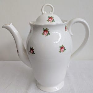 May include: White porcelain teapot with a lid and handle. The teapot is decorated with small pink rose and green leaf floral patterns. The spout has a gold trim. The teapot is designed for serving tea.