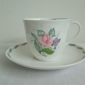 May include: A white teacup and saucer with a floral design. The cup has a pink rose, purple flower, and green leaves. The saucer has a green border.