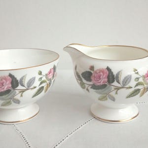 Könnte beinhalten: Ein weißes Porzellan-Zucker- und Milchkännchen-Set mit einem Blumendesign mit rosa Rosen und grünen Blättern. Das Set hat einen goldenen Rand.