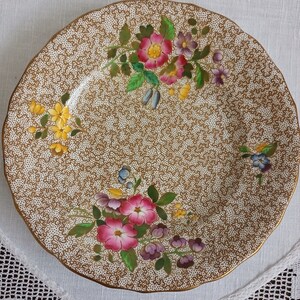 Könnte beinhalten: Ein weißer Keramikteller mit einem goldenen und braunen Blumenmuster. Der Teller zeigt ein zartes Blumenmuster mit rosa, gelben und blauen Blumen.