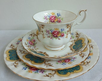 May include: A white teacup and saucer set with a floral pattern and gold trim. The set features a delicate floral design with pink, yellow, and blue flowers. The saucer has a scalloped edge and a teal and gold design.
