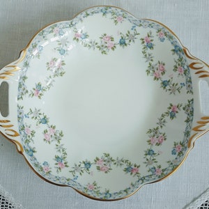 May include: A white porcelain serving dish with gold trim and a floral pattern of pink and blue flowers. The dish has two handles.