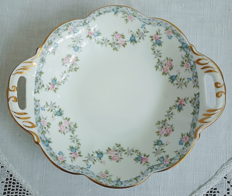 May include: A white porcelain serving dish with gold trim and a floral pattern of pink and blue flowers. The dish has two handles.