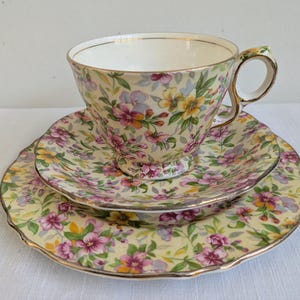 May include: A porcelain teacup and saucer set featuring a pale yellow background with a colourful floral pattern of pink, yellow, and purple flowers with green leaves. The set includes a scalloped edge and gold trim on the rim and handle.