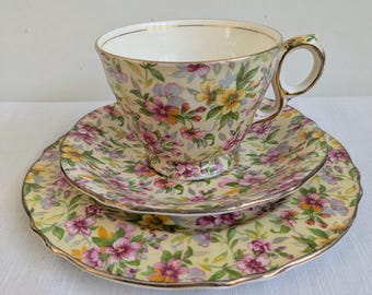 May include: A porcelain teacup and saucer set featuring a pale yellow background with a colorful floral pattern of pink, yellow, and purple flowers with green leaves. The set includes a scalloped edge and gold trim on the rim and handle.