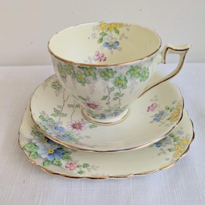 May include: A vintage tea set comprising a teacup, saucer, and side plate. The cream-colored pieces are decorated with a floral design in blue, pink, and yellow. The edges are scalloped and finished with gold trim. The teacup has a curved handle.