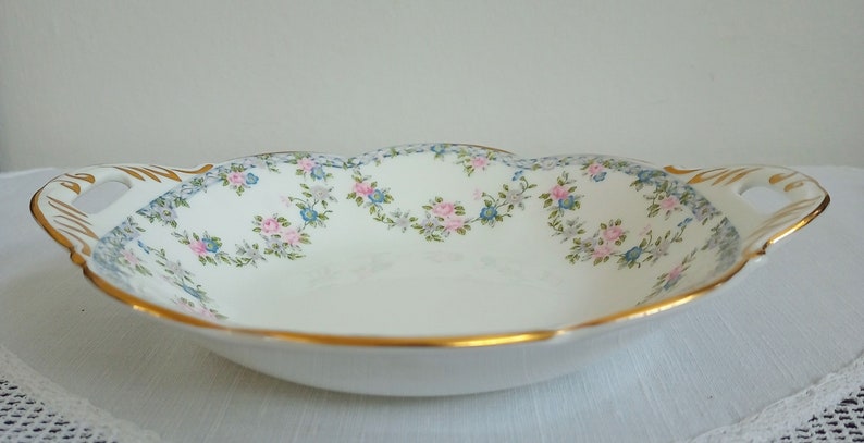 May include: A white porcelain serving dish with a scalloped edge and two handles. The dish is decorated with a floral pattern of pink and blue flowers on a white background. The rim of the dish is trimmed with a gold band.