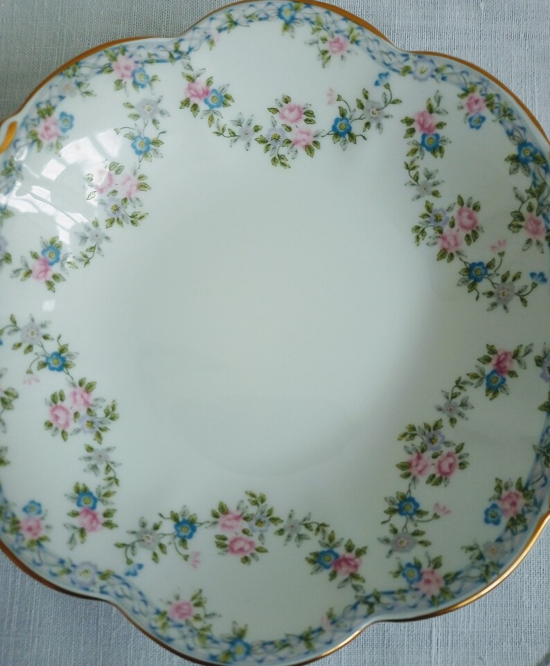 May include: A white porcelain bowl with a scalloped edge and a floral design. The bowl is decorated with pink and blue flowers and green leaves.