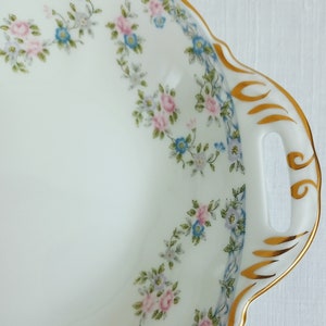 May include: A white porcelain serving dish with a scalloped edge and two handles. The dish is decorated with a floral pattern of pink and blue flowers and green leaves. The rim of the dish is trimmed with a gold band.