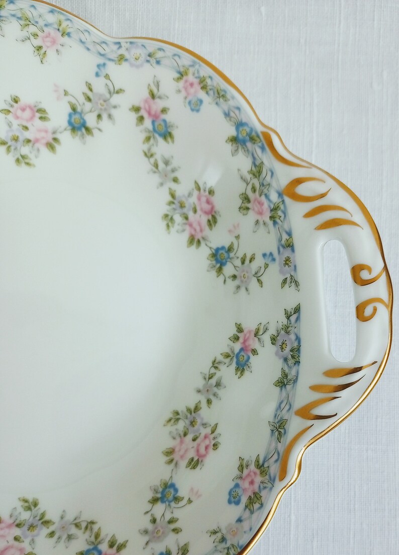 May include: A white porcelain serving dish with a scalloped edge and two handles. The dish is decorated with a floral pattern of pink and blue flowers and green leaves. The rim of the dish is trimmed with a gold band.
